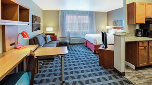 Een compacte hotelkamer met een bed, een bureau met lamp, een sofa, een kleine kitchenette en een raam met gordijnen.