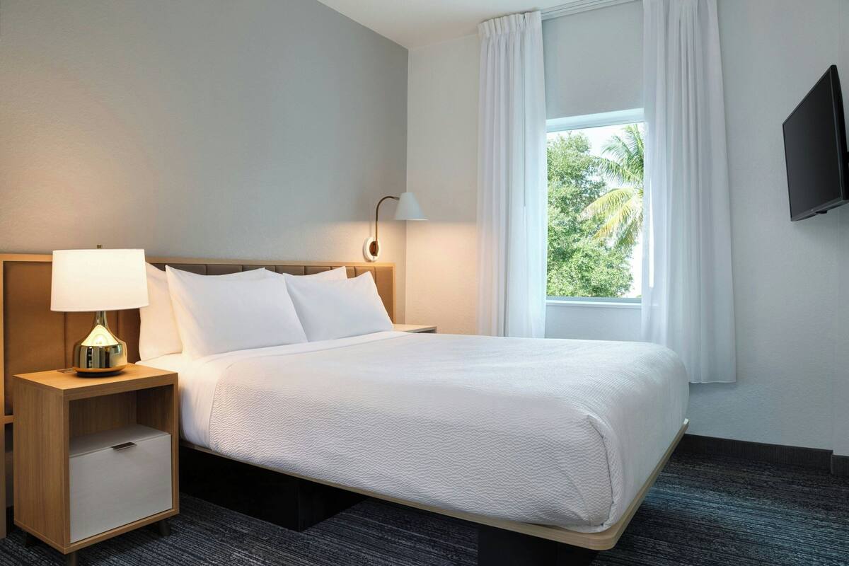 Ein Hotelzimmer mit einem Bett, einem Nachttisch mit Lampe, einem an der Wand montierten Fernseher und einem Fenster mit Vorhängen.