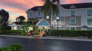 A hotel with a modern design, featuring a central entrance with a blue roof, multiple windows, and a well-maintained garden with palm trees and shrubs.