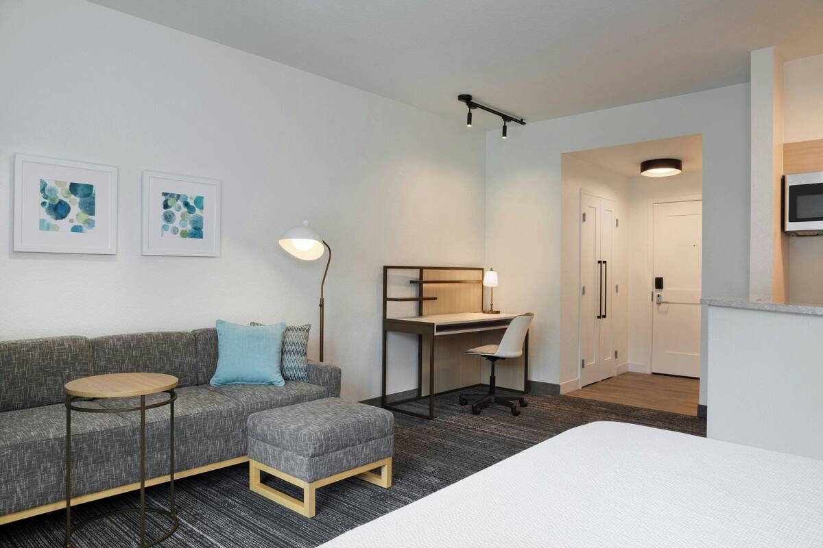 Ein modernes Hotelzimmer mit grauem Sofa, einem kleinen runden Tisch, einem Schreibtisch mit Stuhl, einem Bett, einem Fernseher und zwei gerahmten abstrakten Kunstwerken.