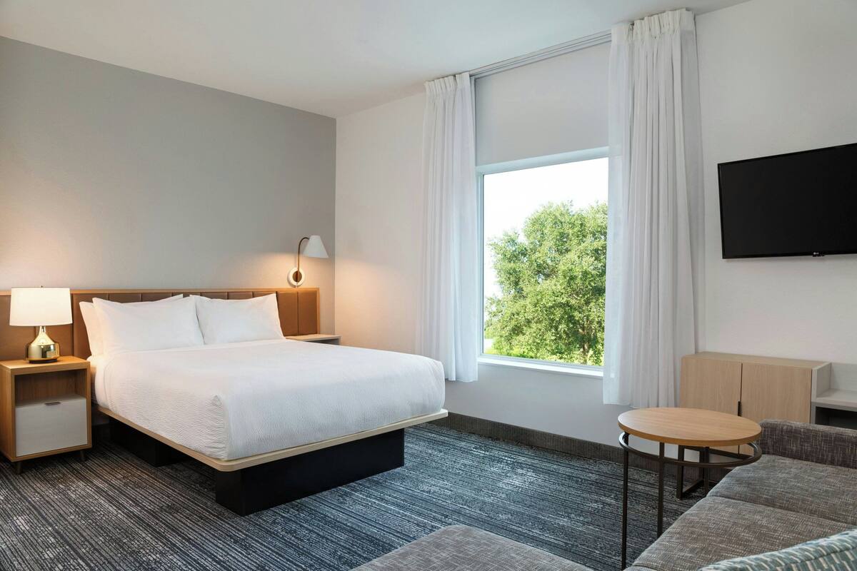 Ein Hotelzimmer mit einem großen Bett, einem Nachttisch mit Lampe, einem kleinen runden Tisch, einer Couch, einem an der Wand montierten Fernseher und einem Fenster mit Vorhängen.