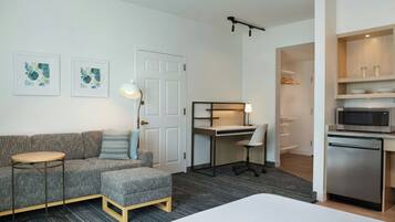 Una habitación de hotel moderna con un sofá gris, una pequeña mesa redonda, un escritorio con una silla, una cama y una kitchenette.
