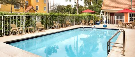 A swimming pool with lounge chairs and umbrellas, surrounded by a fence and palm trees.