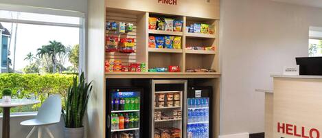 A hotel reception area with a 'PINCH' sign above snack shelves stocked with various snacks and beverages, a 'HELLO Fort Lauderdale' sign, and a seating area with a potted plant.