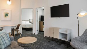 A modern hotel room with a bed, a desk, a chair, a round table, a TV, and a lamp.