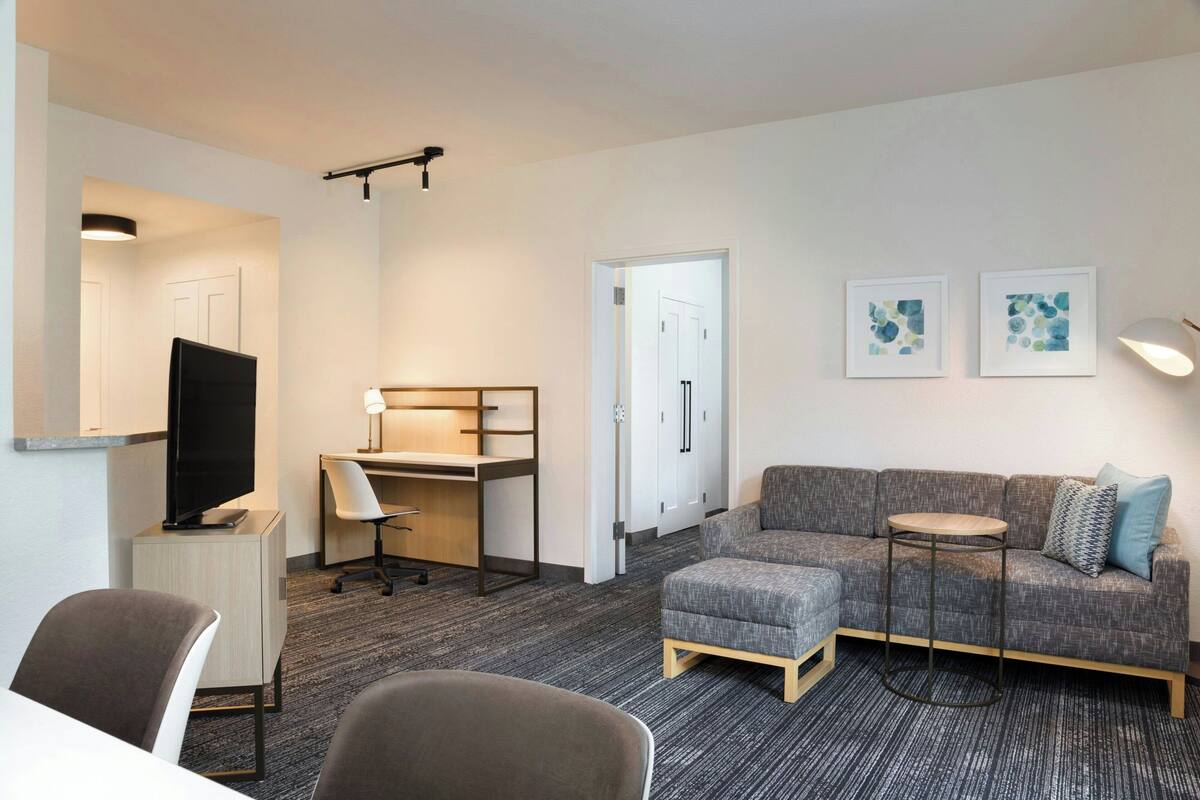 Ein modernes Hotelzimmer mit einem grauen Sofa, einem kleinen runden Tisch, einem Schreibtisch mit Stuhl, einem Fernseher und zwei gerahmten Kunstwerken an der Wand.