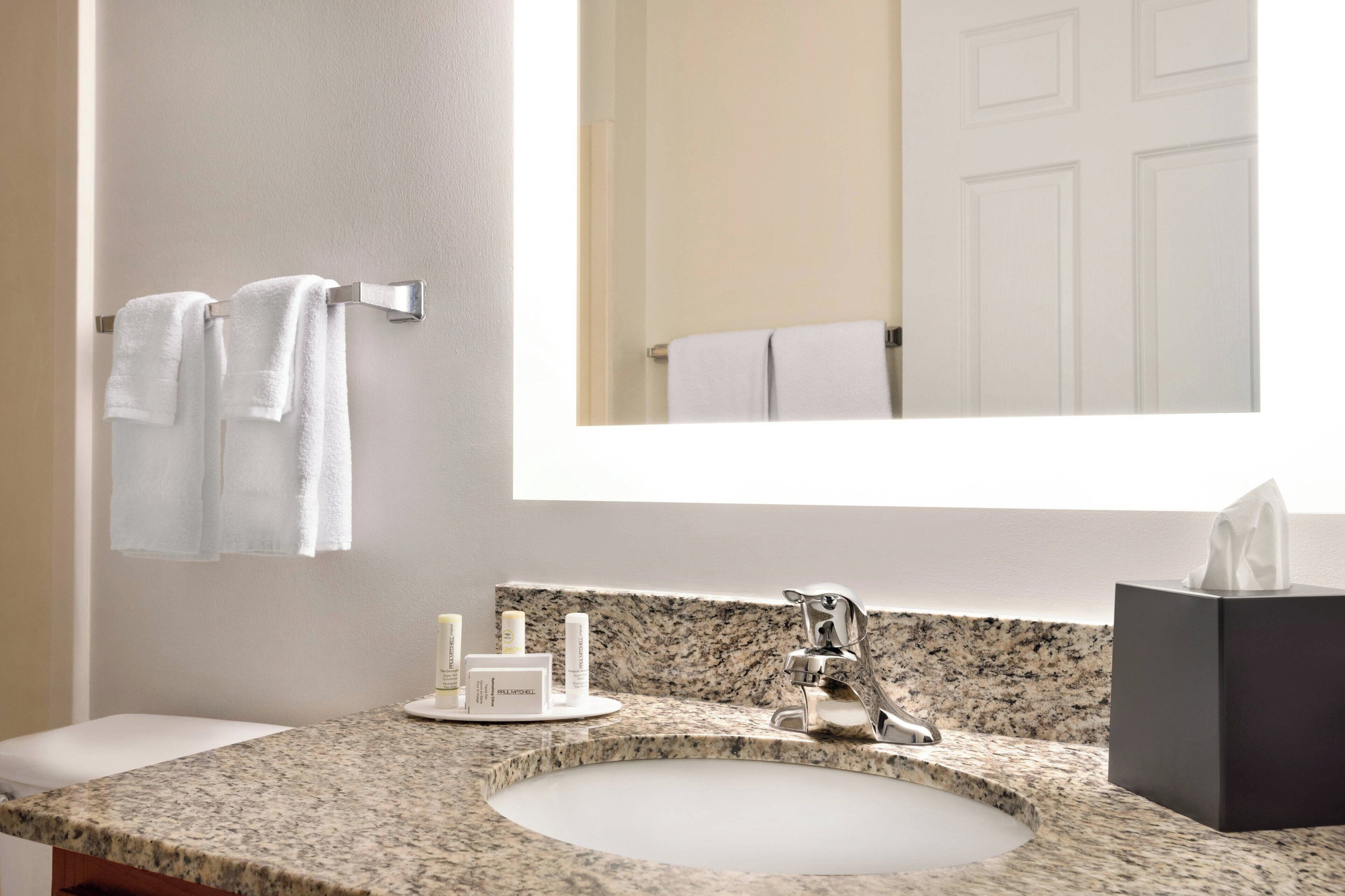 Une salle de bain avec un lavabo doté d’un plan de travail en granit, un miroir, deux serviettes blanches suspendues à un porte-serviettes et une boîte de mouchoirs posée sur le plan de travail.