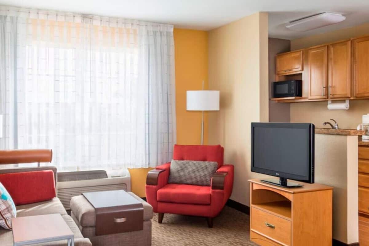 A hotel room with a sofa, armchair, TV, and a small kitchenette.