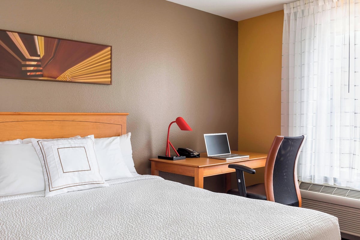 A hotel room with a bed, a desk with a laptop, a chair, a red lamp, and a large abstract painting on the wall.