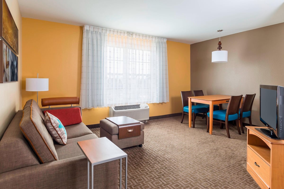 A hotel room with a sofa, a small table, a TV, a dining table with chairs, a window with curtains, and an air conditioning unit.