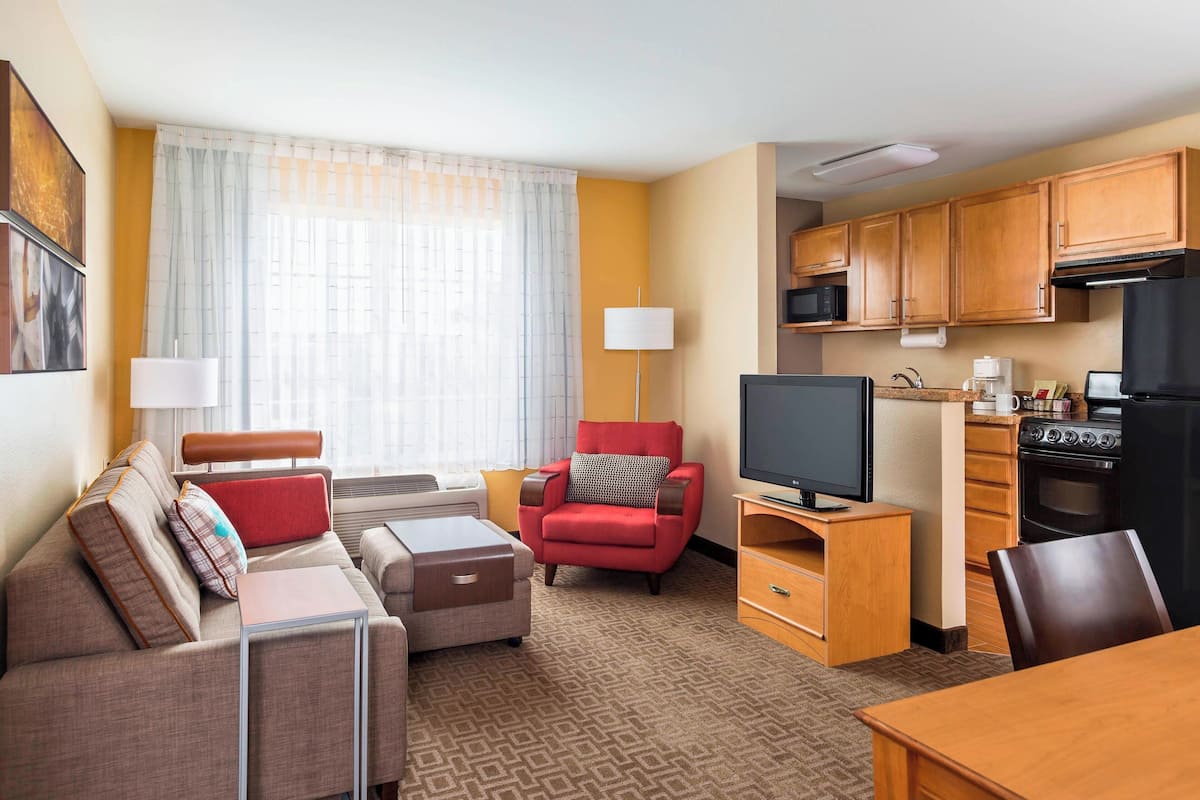 A hotel room with a sofa, armchair, TV, kitchenette, and a large window with curtains.