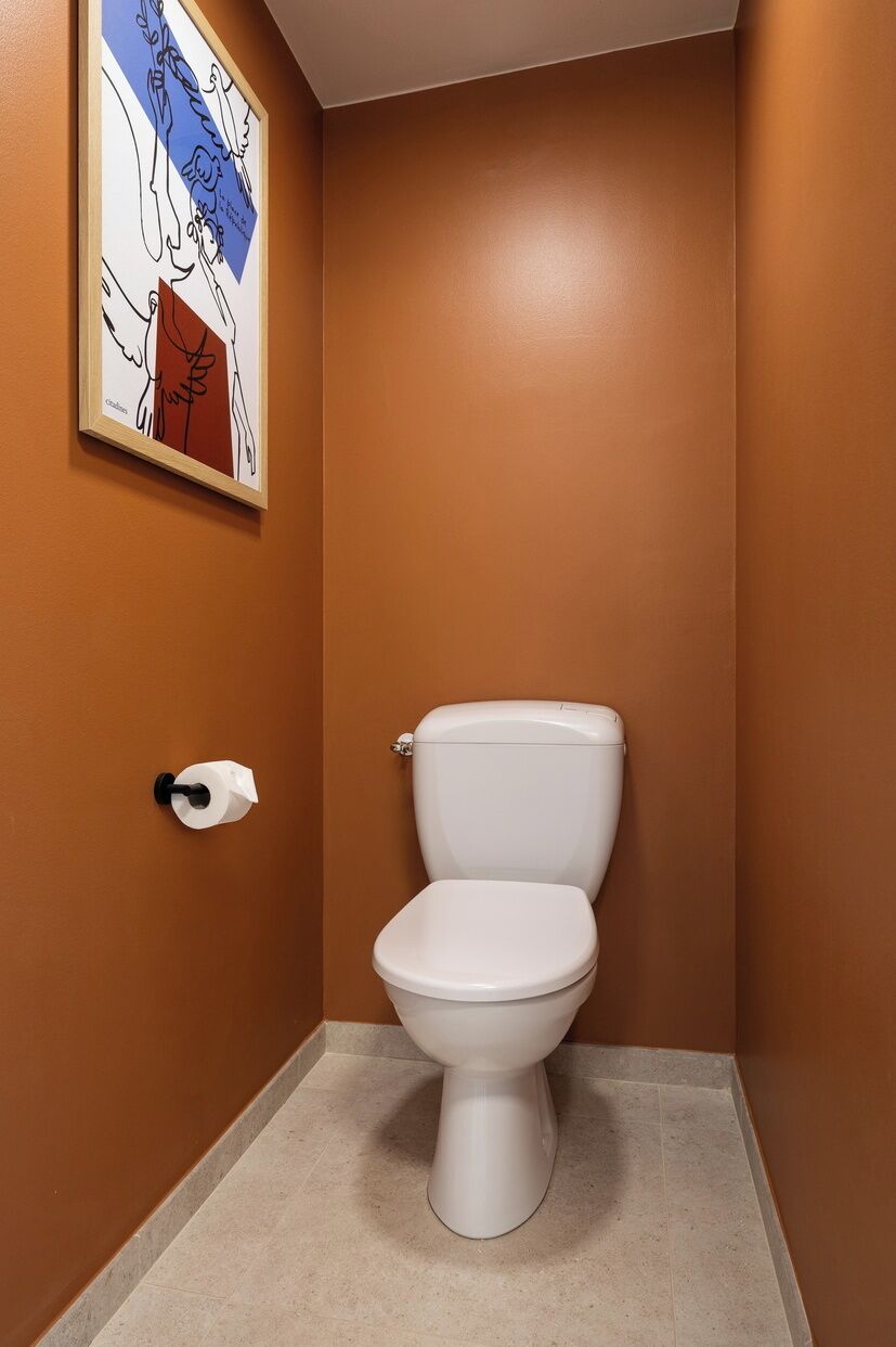 Un baño con inodoro blanco, paredes marrones y una obra de arte enmarcada sobre el inodoro.
