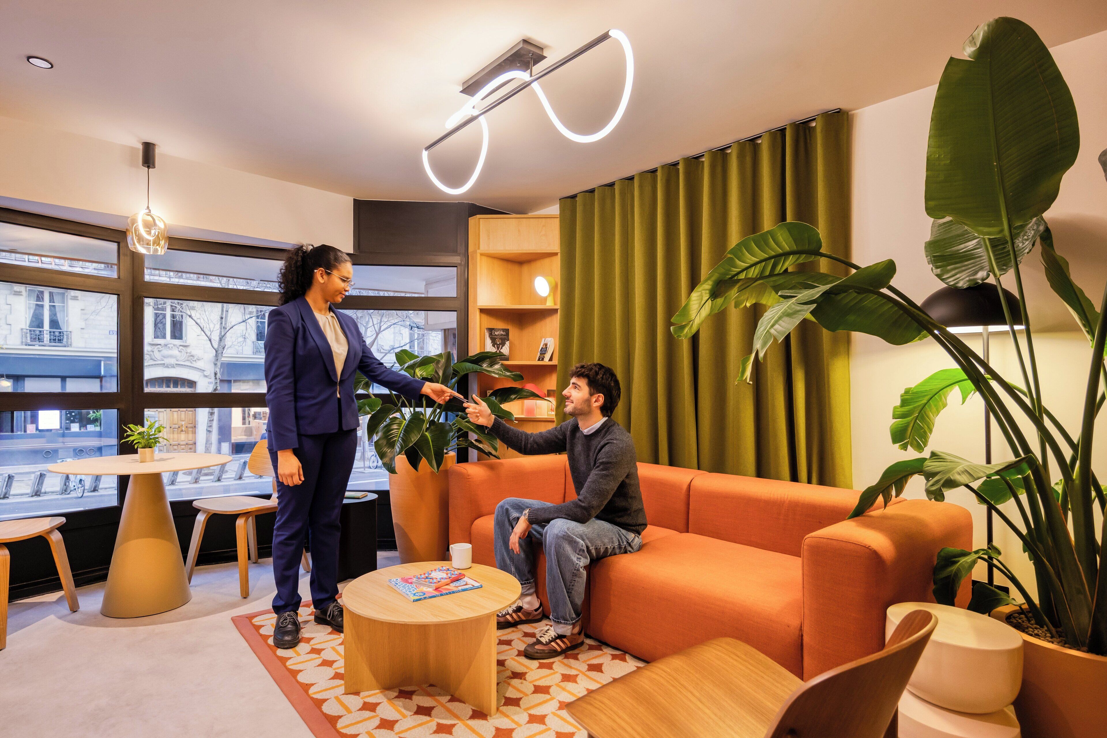 A modern office space with a large window, a round wooden table, an orange sofa, and several potted plants.