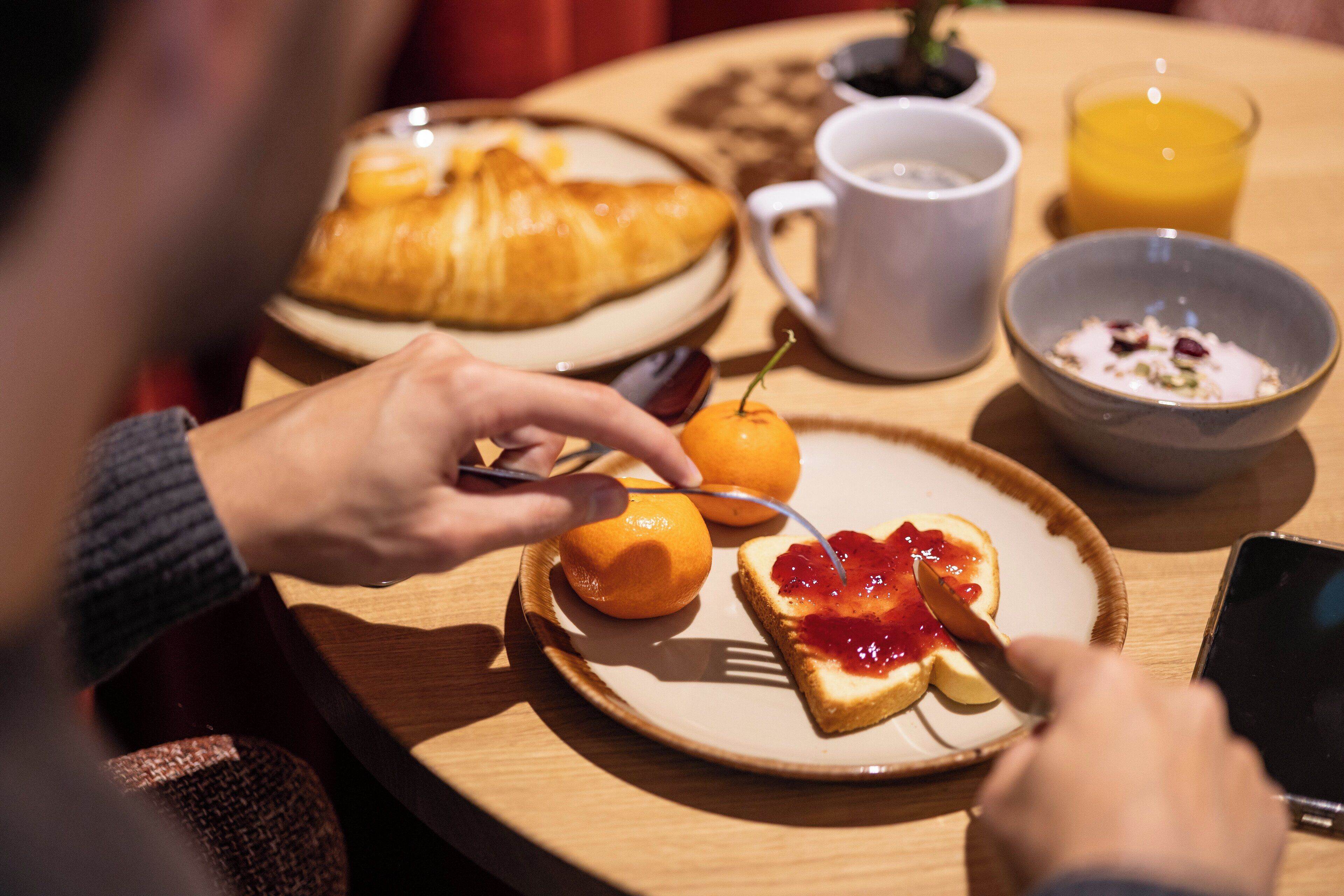 A breakfast setting with a croissant, orange, toast with jam, a mug, a bowl of cereal, and a glass of orange juice on a wooden table.