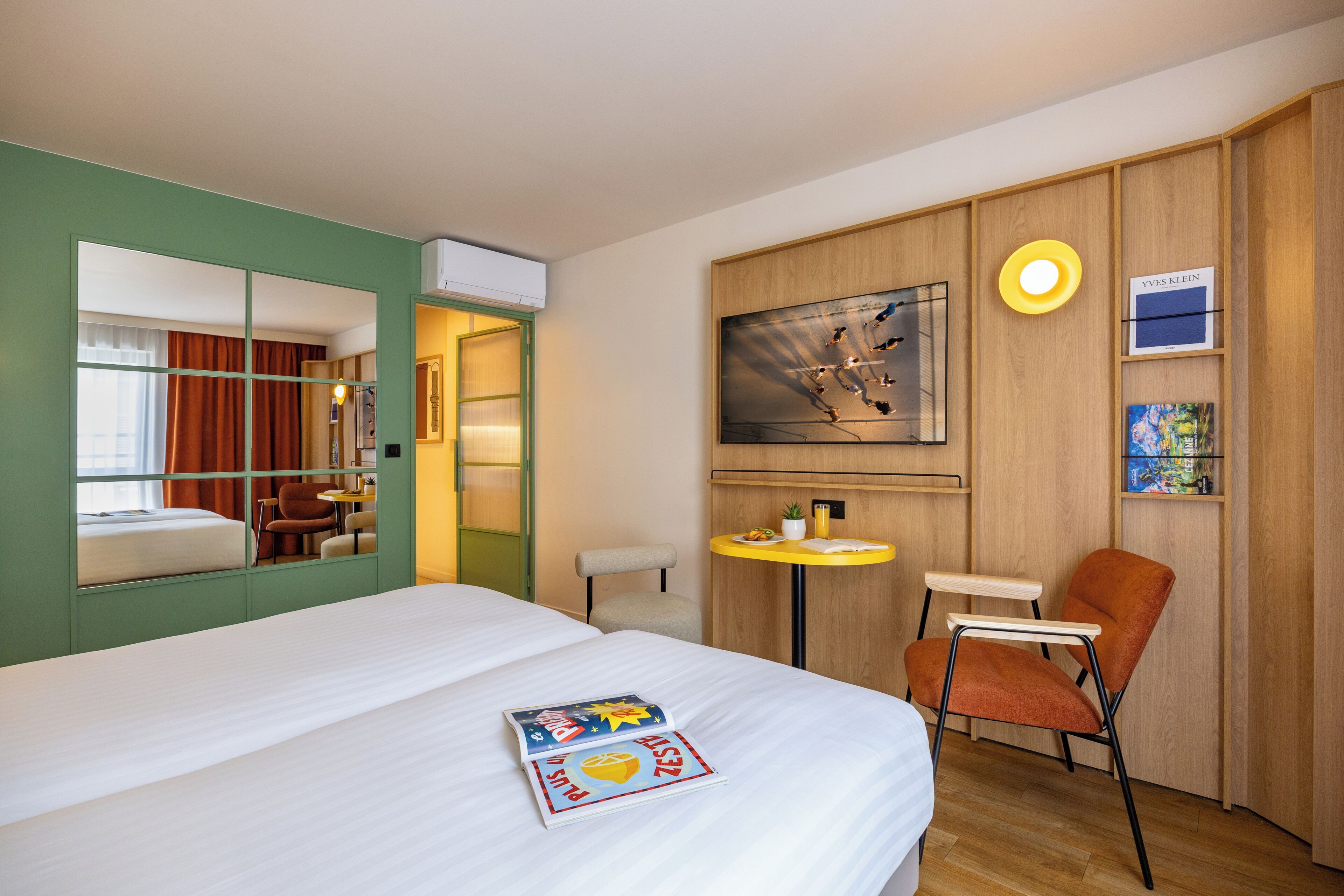 Een hotelkamer met een bed, een bureau, een stoel, een klein tafeltje, een spiegel, een televisie en kunst aan de muur.