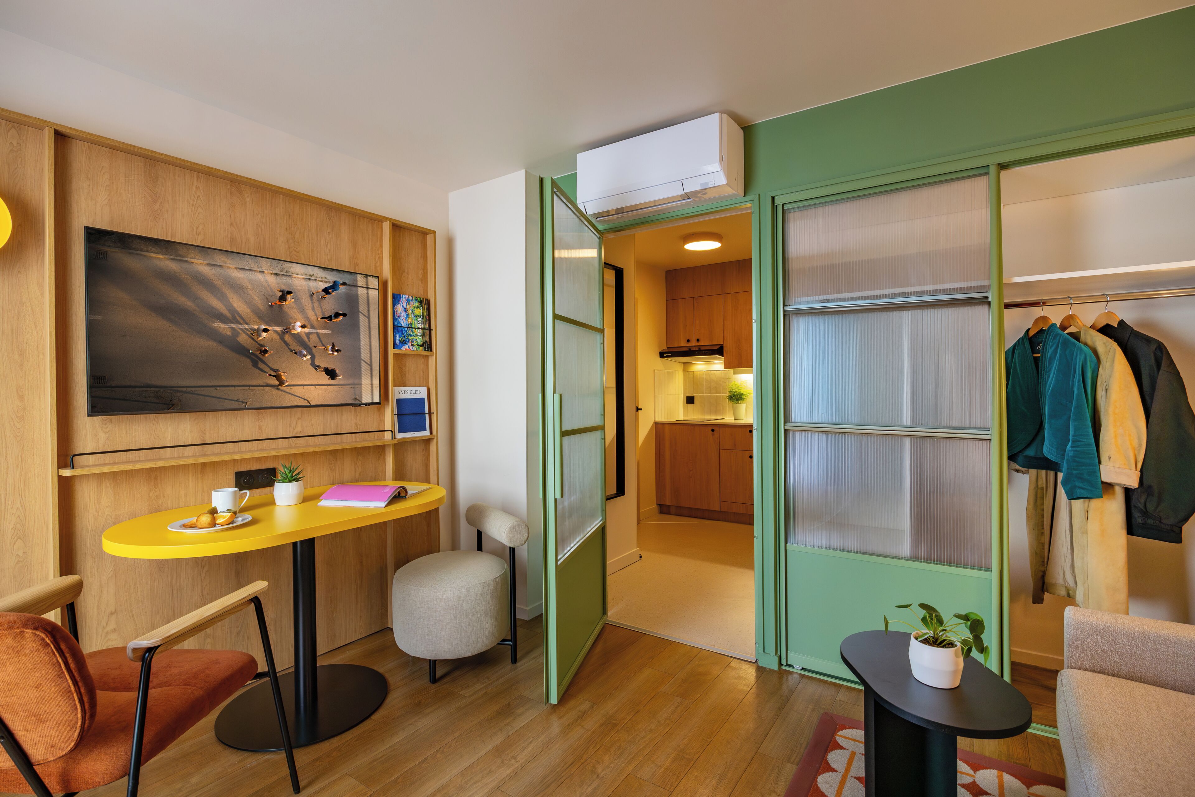 Ein kompaktes Hotelzimmer mit Holzwand, einem kleinen Esstisch, einer Couch, einem Sessel und einem Kleiderständer mit Kleidung.