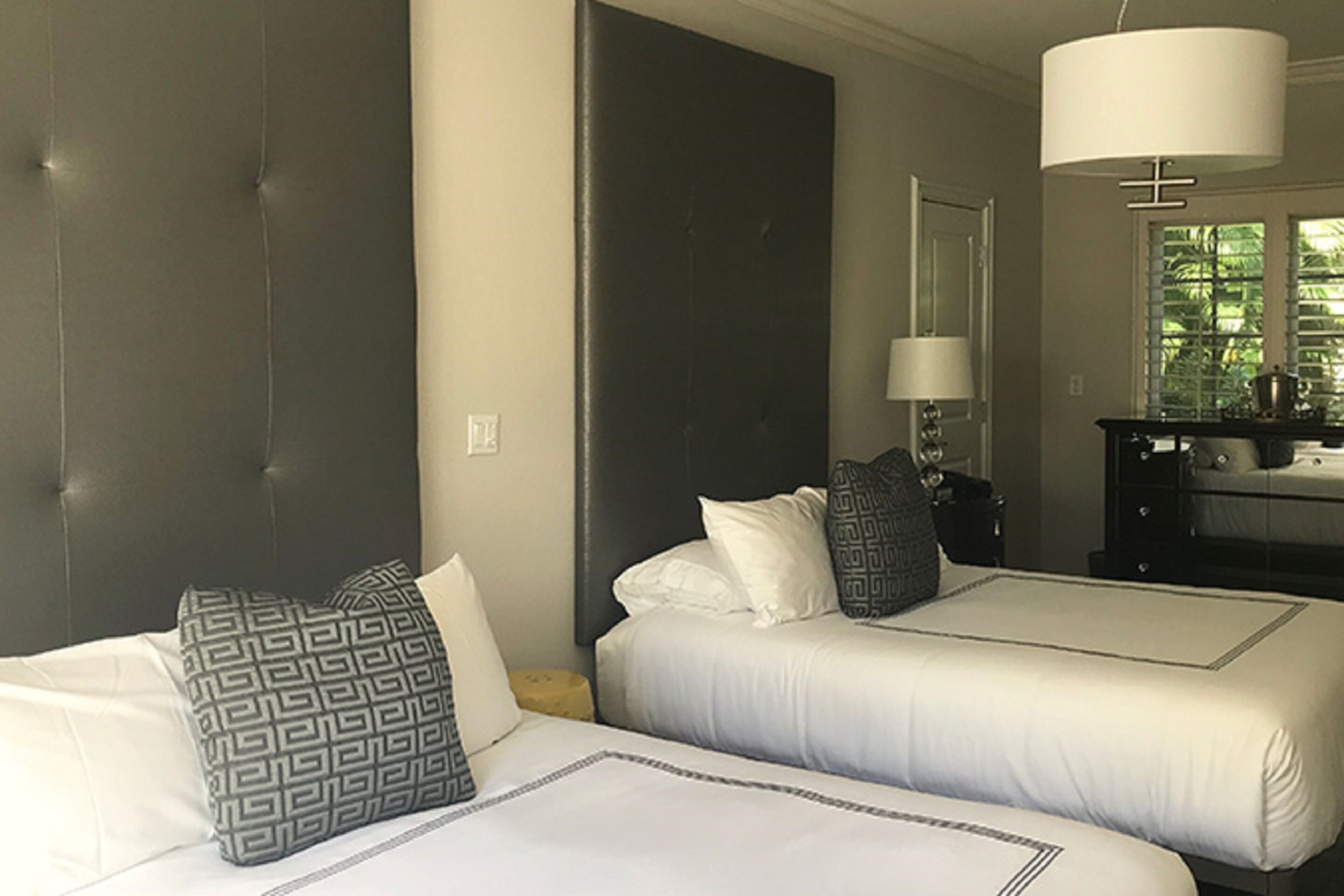 Une chambre d’hôtel avec deux lits, une grande tête de lit sombre, une table de chevet avec une lampe et une fenêtre avec des stores.