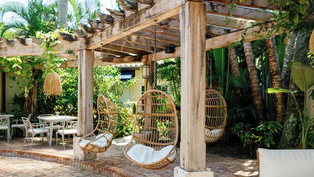 Un espace extérieur aménagé, abrité et doté de chaises en osier et d’un canapé blanc, entouré d’une végétation luxuriante et de palmiers.