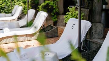 Une rangée de chaises longues blanches, chacune agrémentée d’une serviette et d’une bouteille d’eau, est disposée sur une terrasse en briques, avec une végétation luxuriante en arrière-plan.