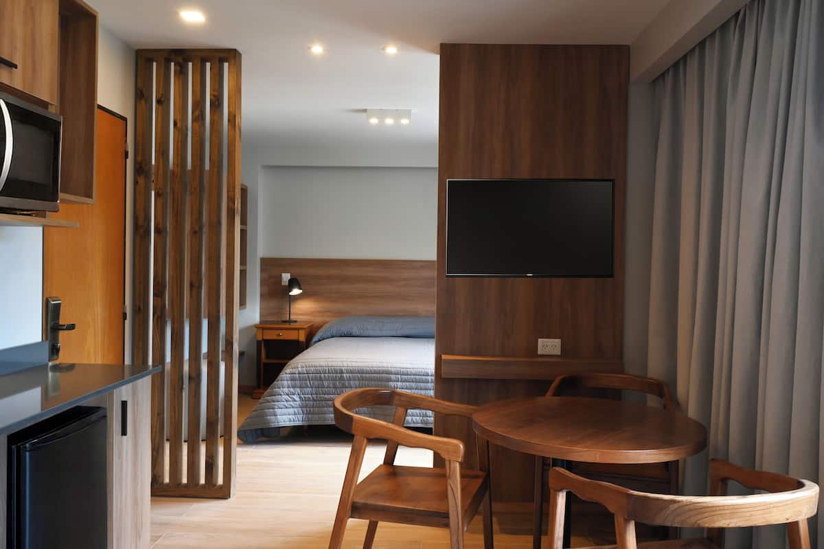 Een moderne hotelkamer met een bed, een televisie, een eettafel met stoelen, een houten scheidingswand en een bureau met een computer.