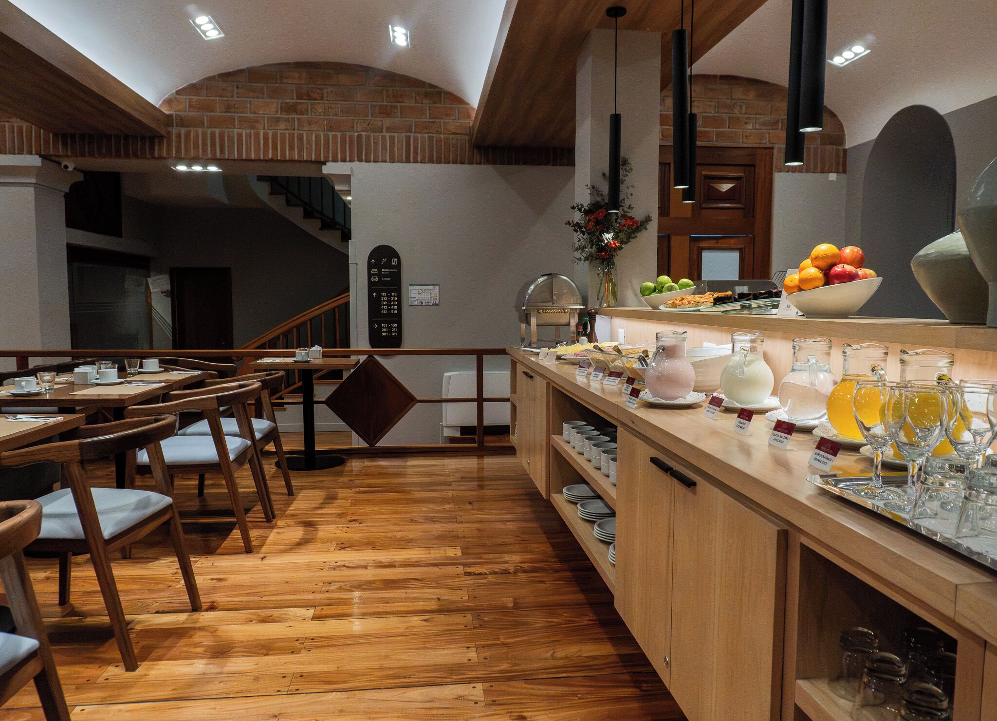 A modern kitchen with wooden flooring, a central island with a fruit bowl, and a staircase leading to an upper level.