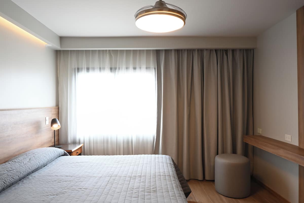 Een slaapkamer met een bed, een bedlampje, een raam met gordijnen en een rond krukje.