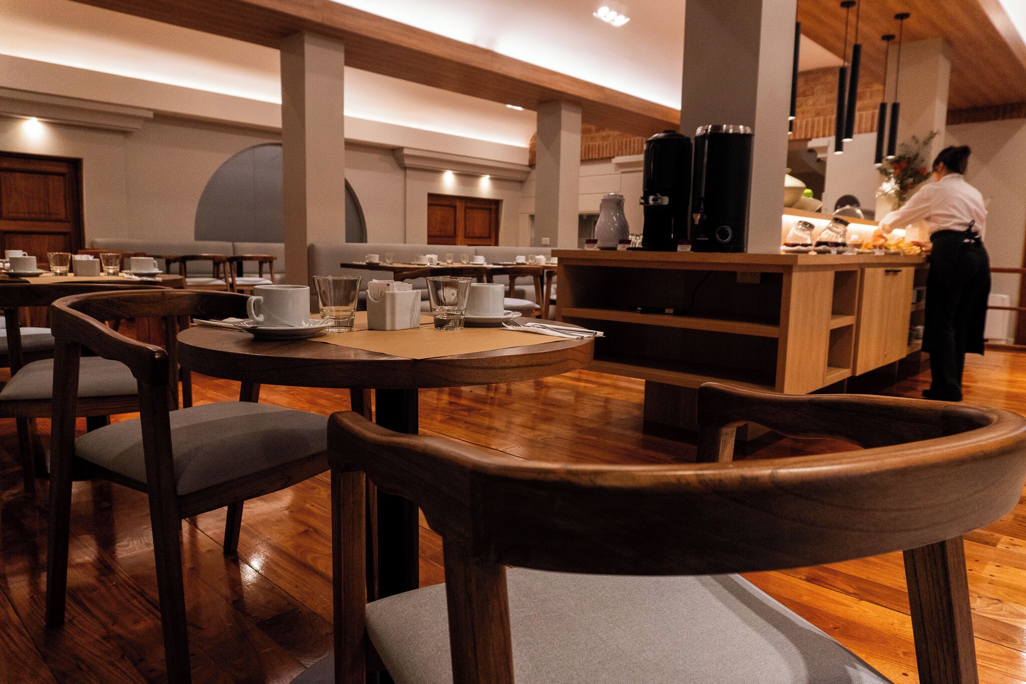 A restaurant with round tables and chairs, a bar area, and a person working at the bar.