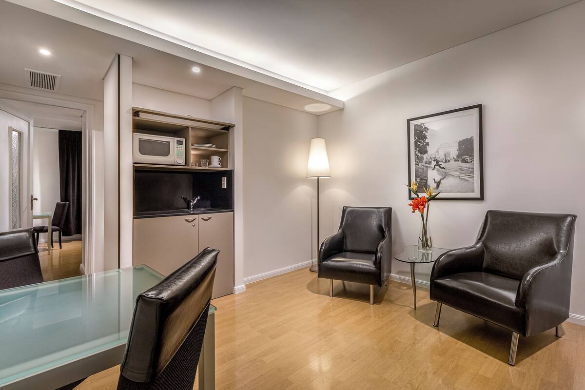A modern hotel room with a glass dining table, two leather armchairs, a standing lamp, a microwave, and a framed picture on the wall.