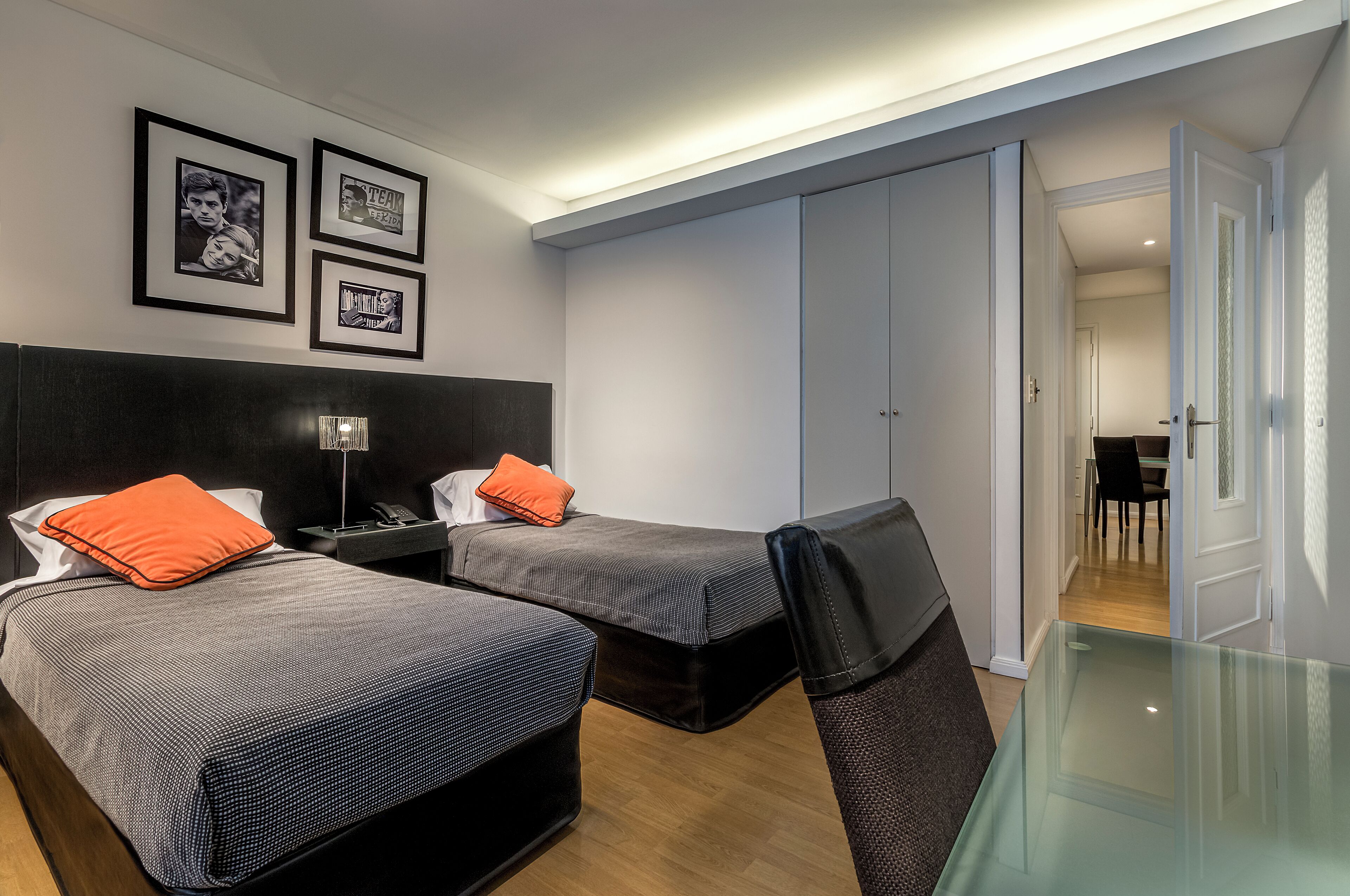 A hotel room with two beds, a desk, a chair, and a glass table. There are framed pictures on the wall and a wardrobe.