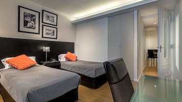 A hotel room with two beds, a desk, a chair, and a glass table. There are framed pictures on the wall and a wardrobe.