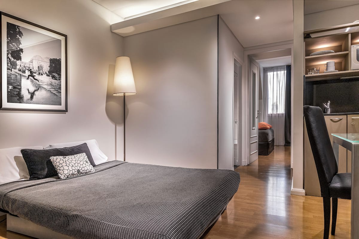 A modern hotel room with a large bed, a bedside table, a lamp, a framed picture on the wall, and a view into the adjacent room.