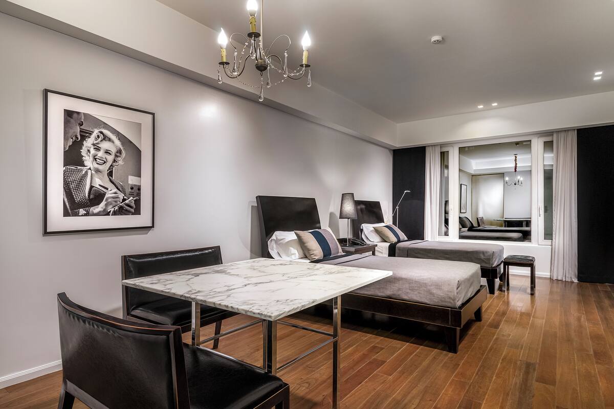 A modern hotel room with a marble table, leather chairs, a bed with a grey mattress, a small bedside table with a lamp, and a framed picture on the wall.