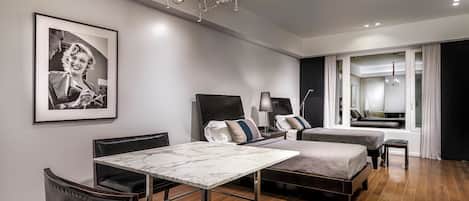 Una habitación de hotel moderna con una mesa de mármol, sillas de cuero, una cama con colchón gris, una mesita de noche con lámpara y un cuadro en la pared.