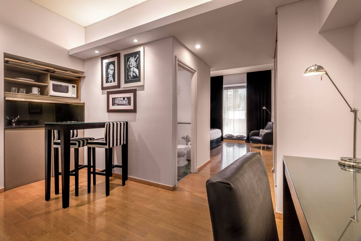 A modern living space with a kitchen area, dining table, and seating. There is a visible door leading to a bathroom.