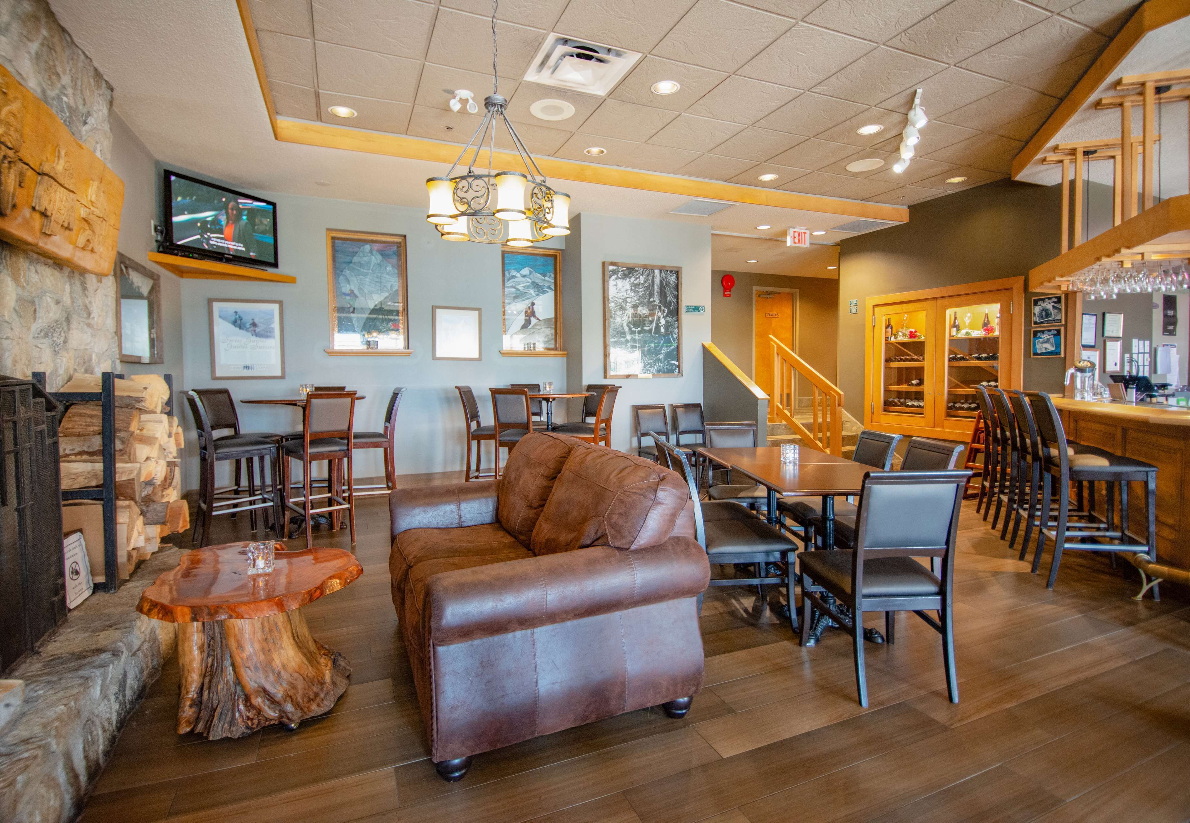 A spacious lobby with a leather sofa, wooden tables, and chairs, a bar area with glasses, and framed pictures on the walls.