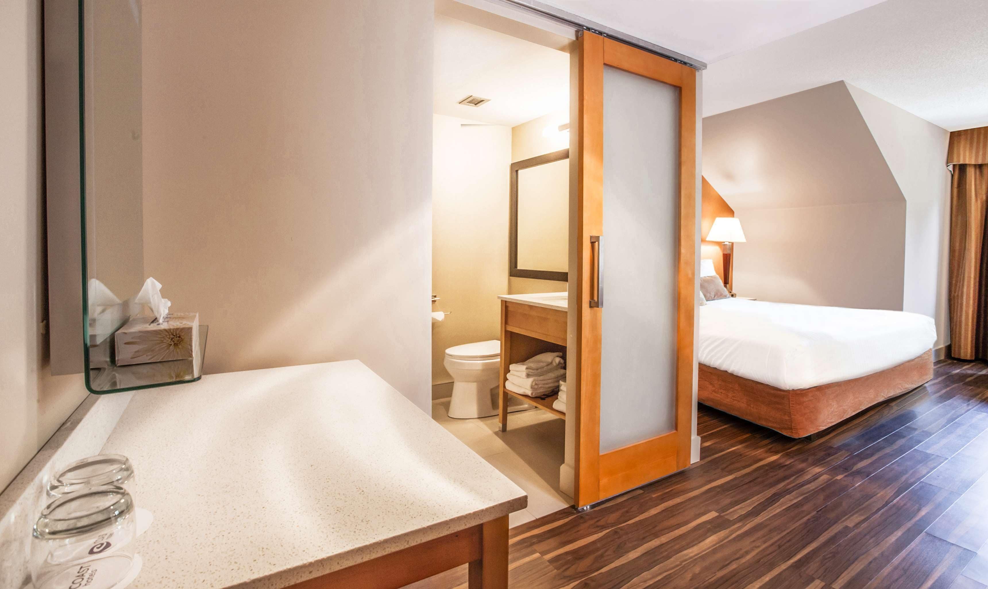 Ein Hotelzimmer mit Holzboden, einem Bett, einem Nachttisch mit Taschentuchbox, einem Spiegel und einem Badezimmer inklusive Toilette und Handtuchhalter.