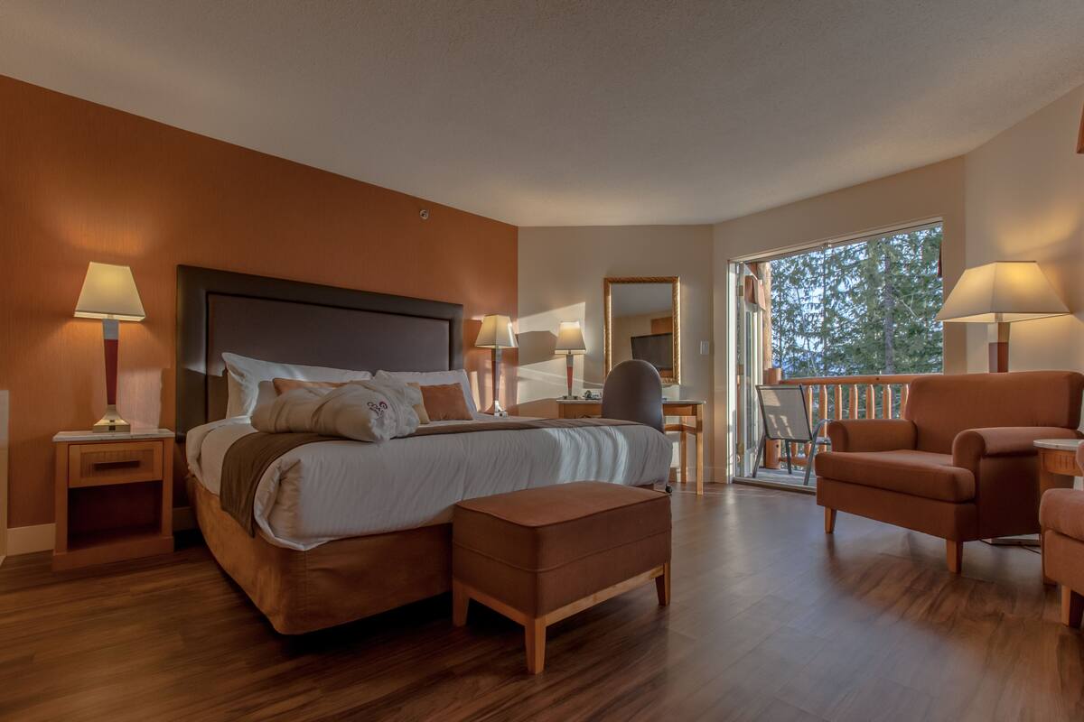 A modern hotel room with a large bed, a sitting area with a chair, a desk, and a balcony with a view.