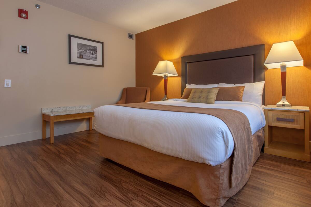 A hotel room with a large bed, two bedside lamps, a nightstand, a bench, and a framed picture on the wall.