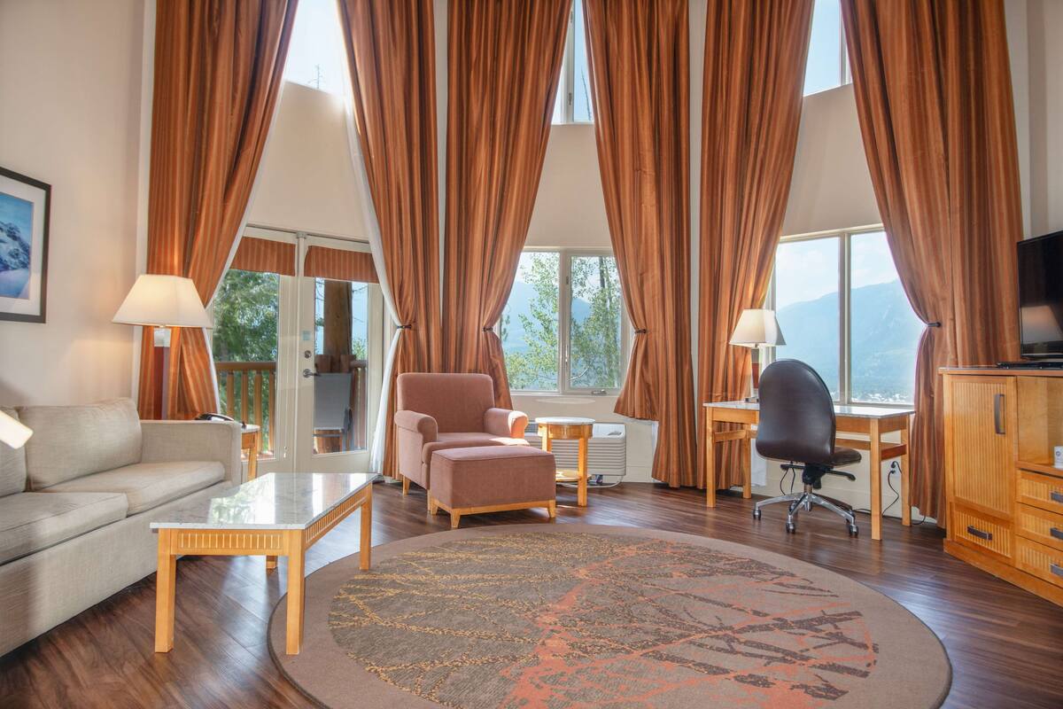 A spacious living room with a sofa, armchair, and coffee table, large windows with curtains, and a view of the outdoors.