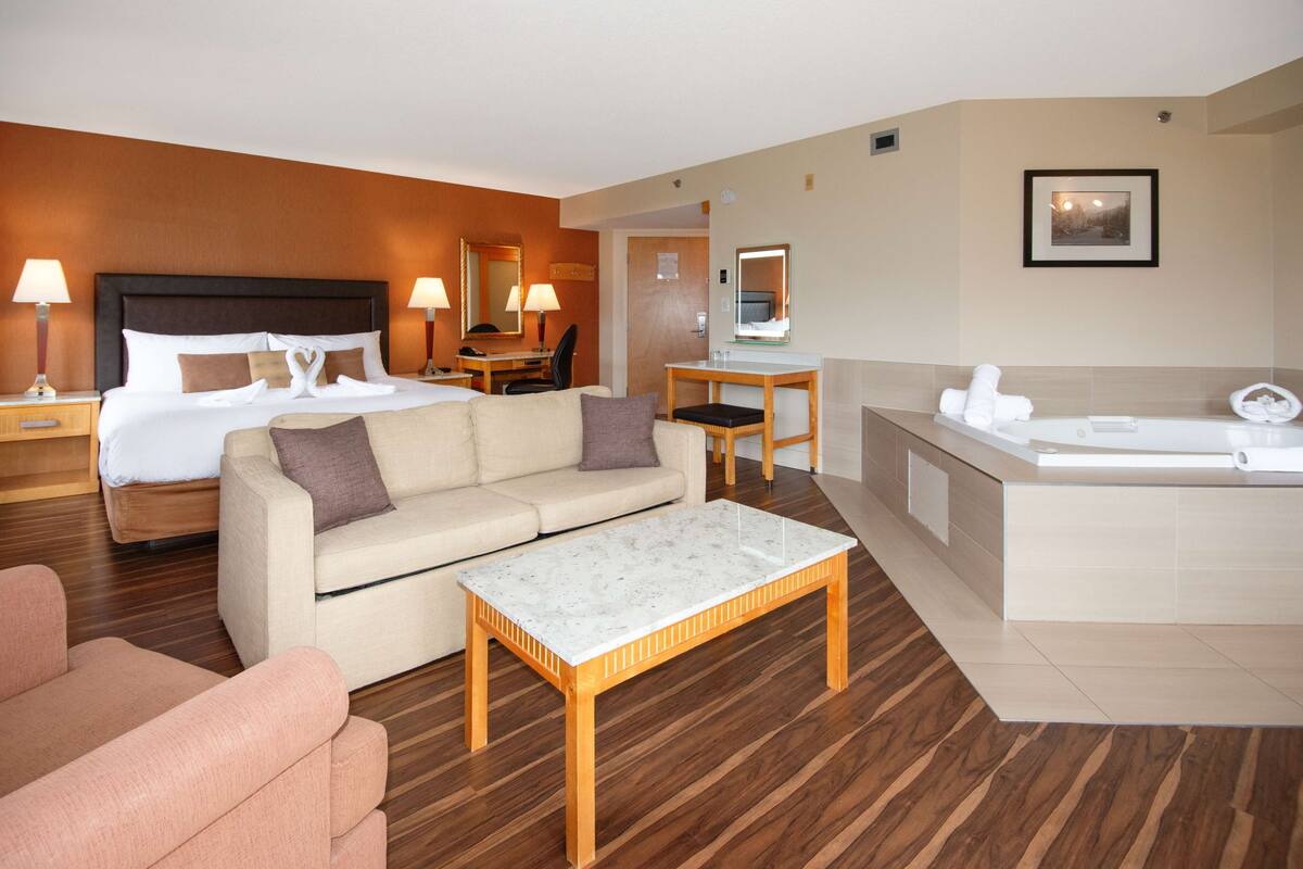 A hotel room with a bed, a sofa, a coffee table, a desk, a chair, a bathroom with a bathtub, and a mirror.