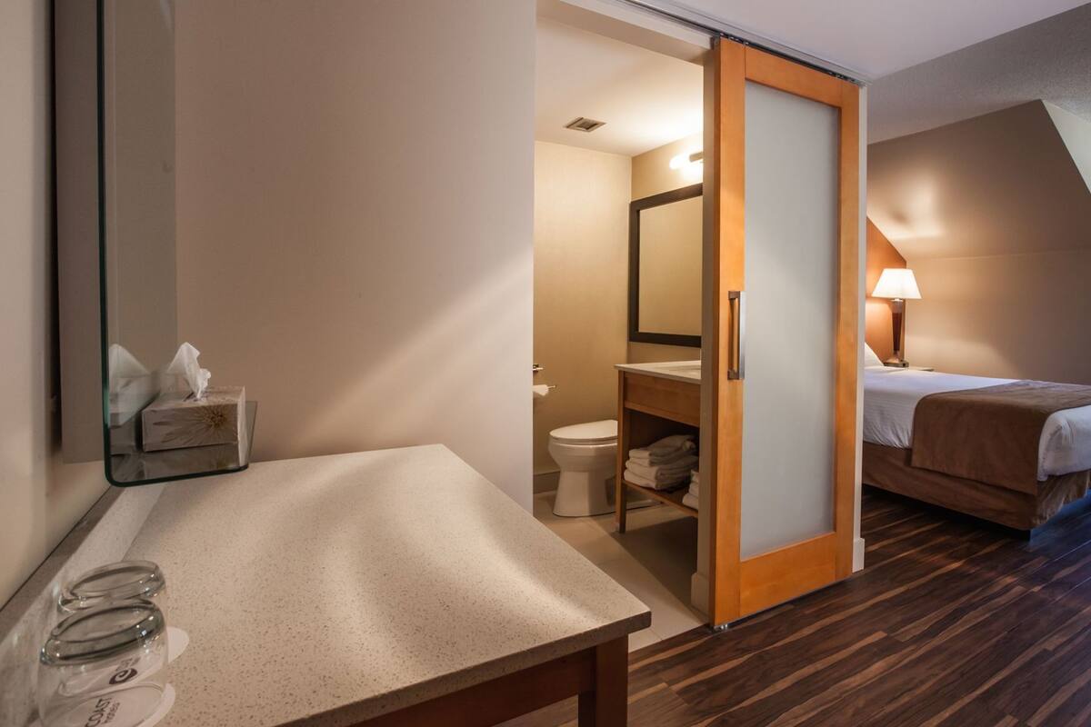 A hotel room with a wooden floor, a bed, a bedside table, a mirror, a bathroom with a toilet, and a shower area.