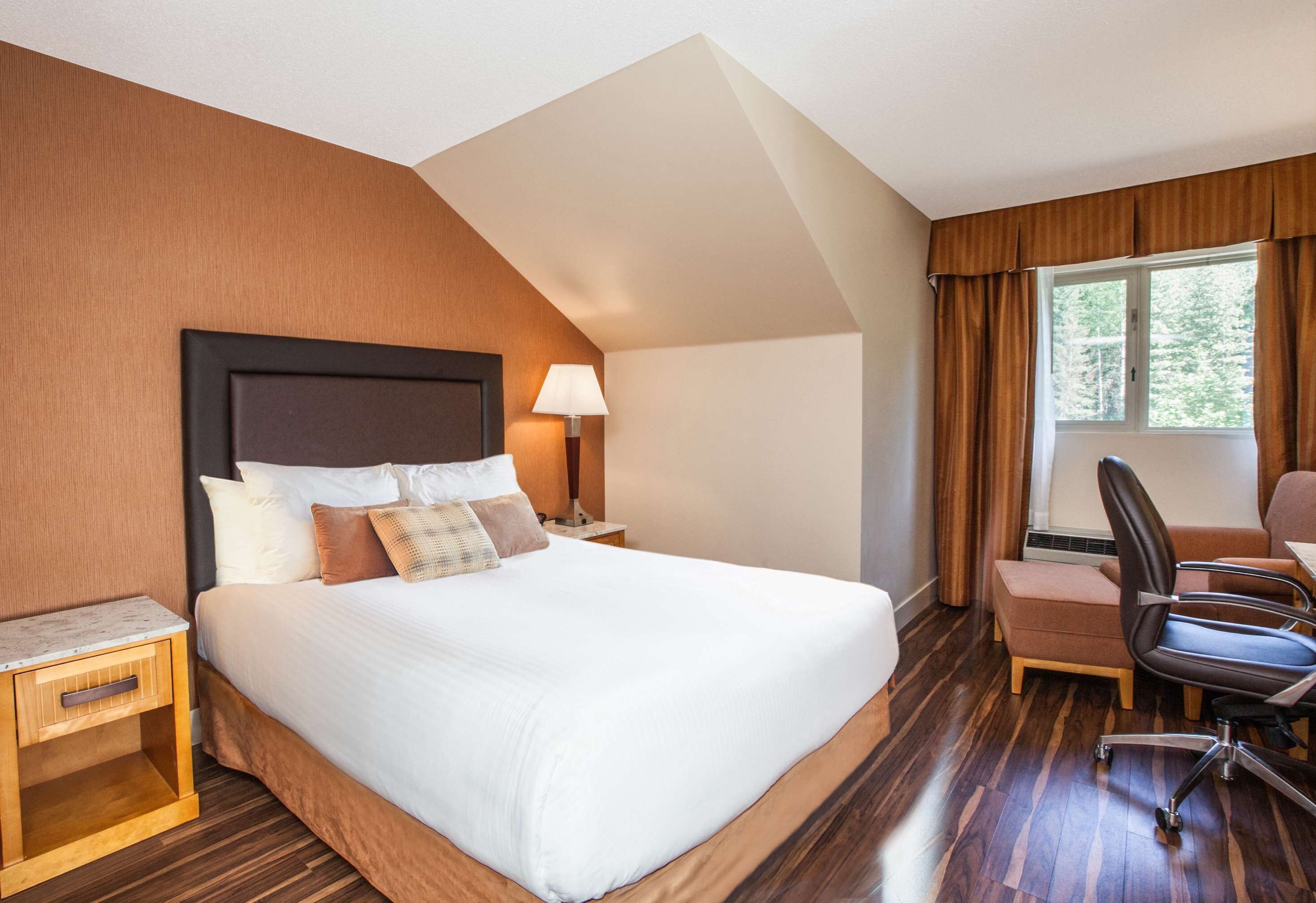 A hotel room with a large bed, a desk with a chair, a window with curtains, and a lamp on the nightstand.