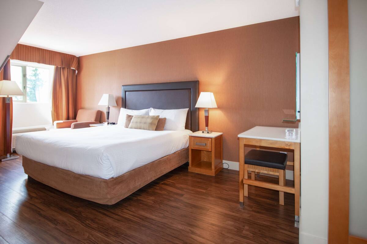 A hotel room with a large bed, two bedside tables with lamps, a small desk with a chair, and a window with curtains.