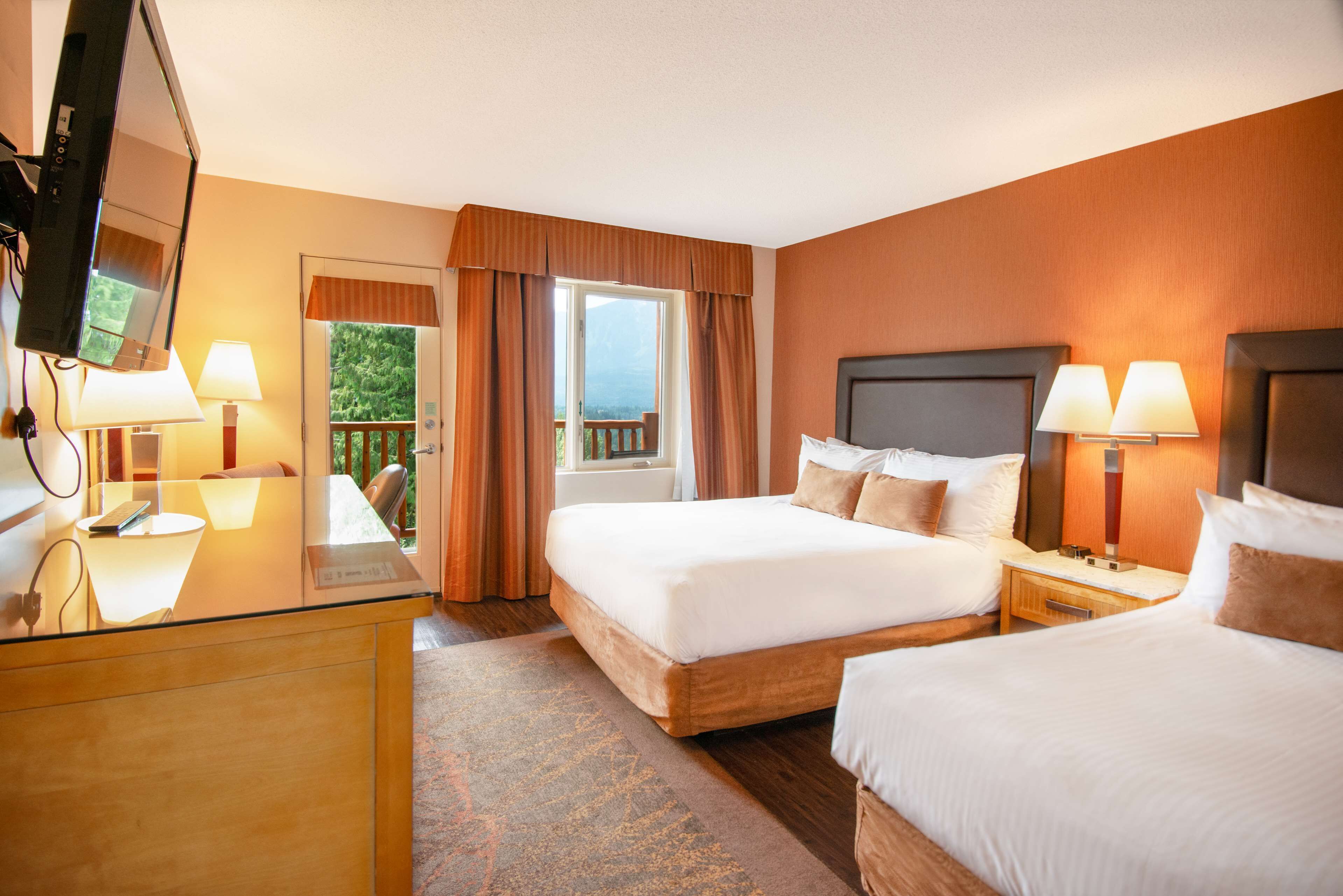Ein Hotelzimmer mit zwei Betten, einem Schreibtisch mit Computer, einem Fernseher und einem Balkon mit Vorhängen.