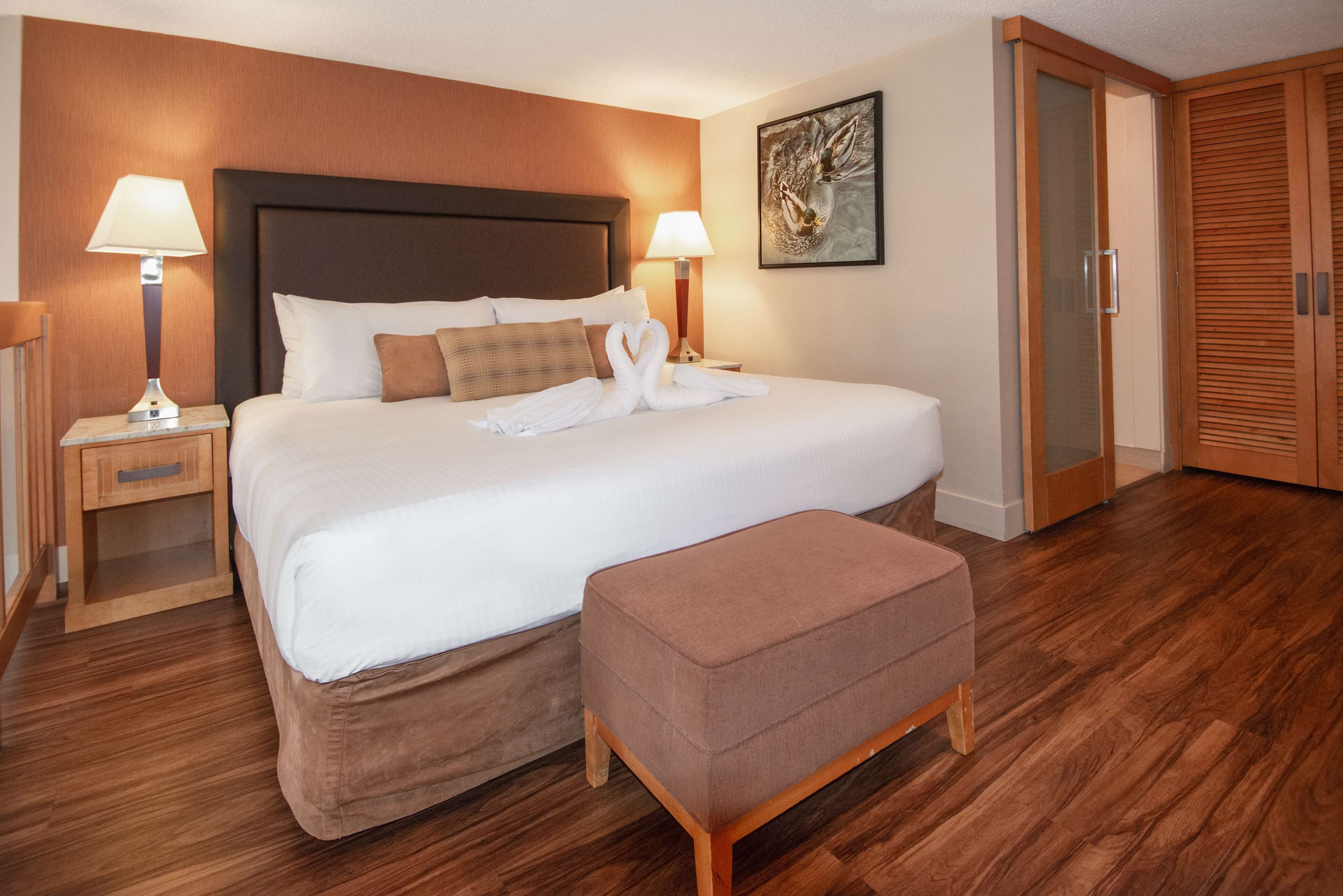 A neatly made bed with a headboard, two bedside lamps, a wooden floor, a wooden bench, and a wooden wardrobe.
