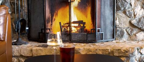 A stone fireplace with a fire, a glass of red beverage, and a wine glass on a table.