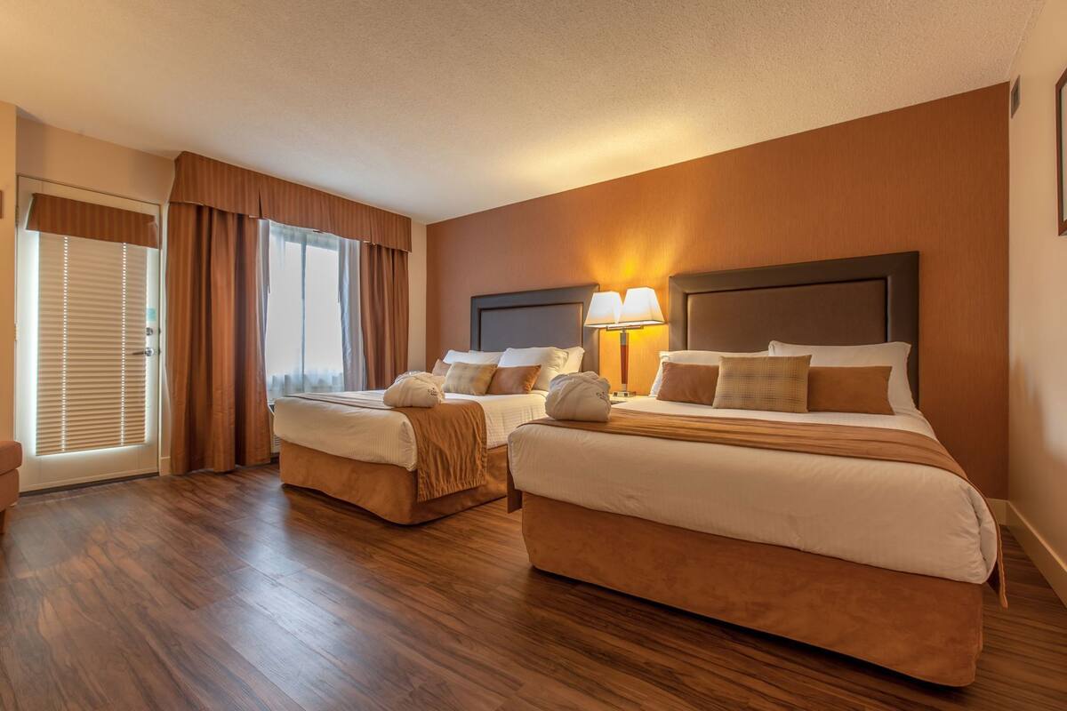 A hotel room with two beds, a large window with curtains, a door with blinds, and a nightstand with a lamp.