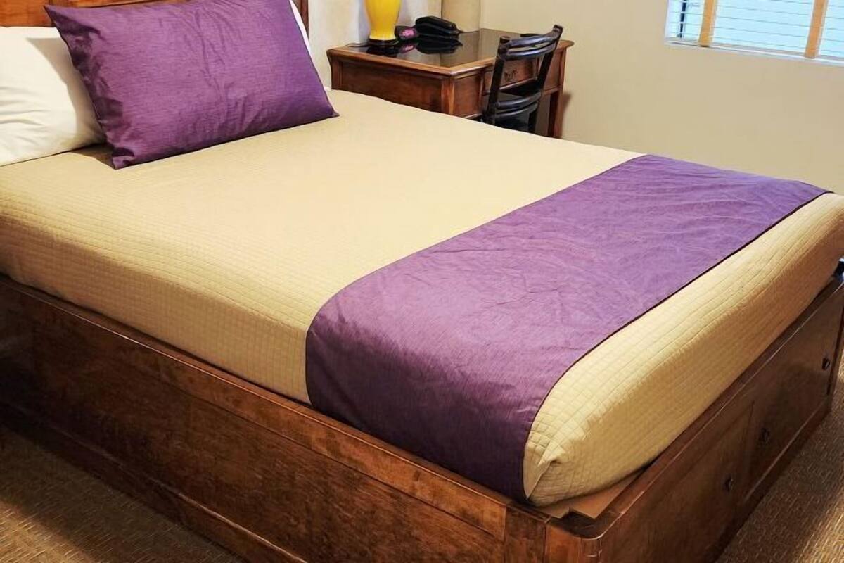 Una cama con estructura de madera, colchón color crema y manta morada.