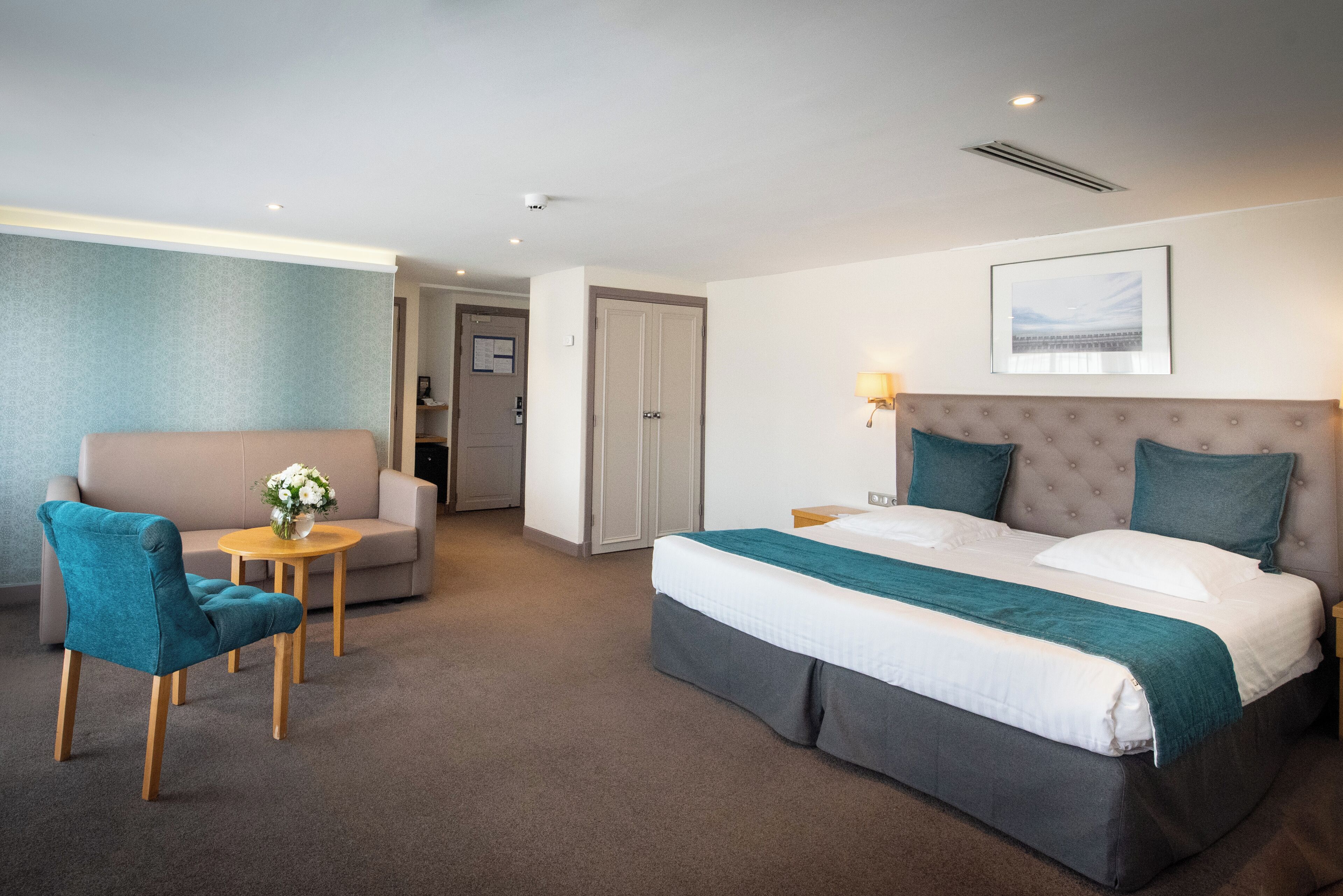 A hotel room with a bed, a sofa, a chair, a small table with a vase, and a framed picture on the wall.