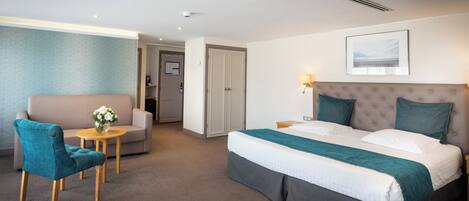 Una habitación de hotel con cama, sofá, sillón, una mesita con un florero y un cuadro en la pared.
